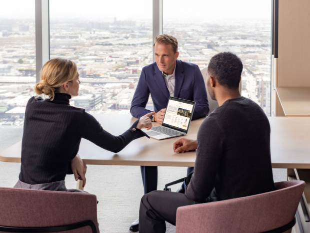 Three people at a conference room table discussing what is displayed on a laptop screen.